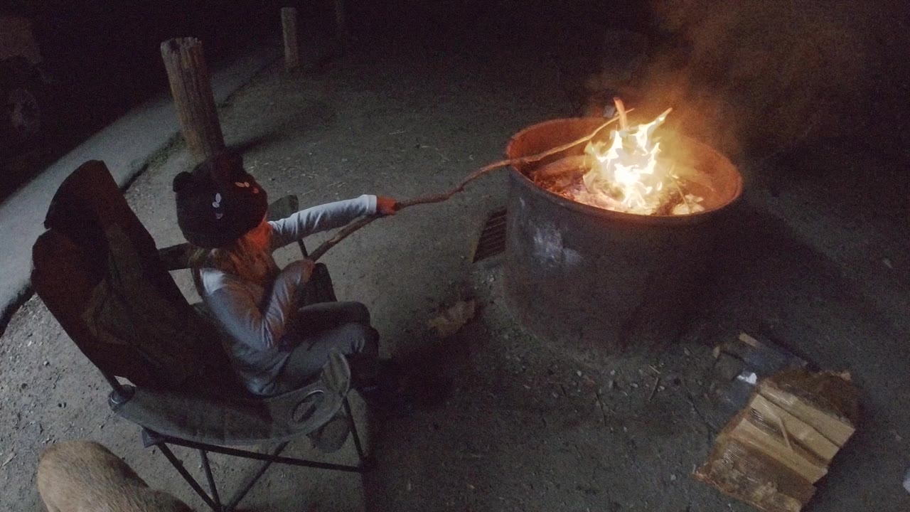 High-angle view in San Clemente, California, showing a child seated in a folding chair while holding a long stick into a bright round fire pit at night.