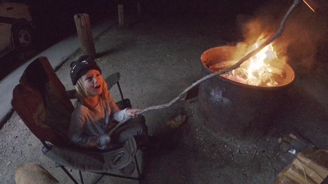 At 0:00 in San Clemente, California, a young child sits in a folding chair and extends a long stick toward a round fire pit with flames on the right.