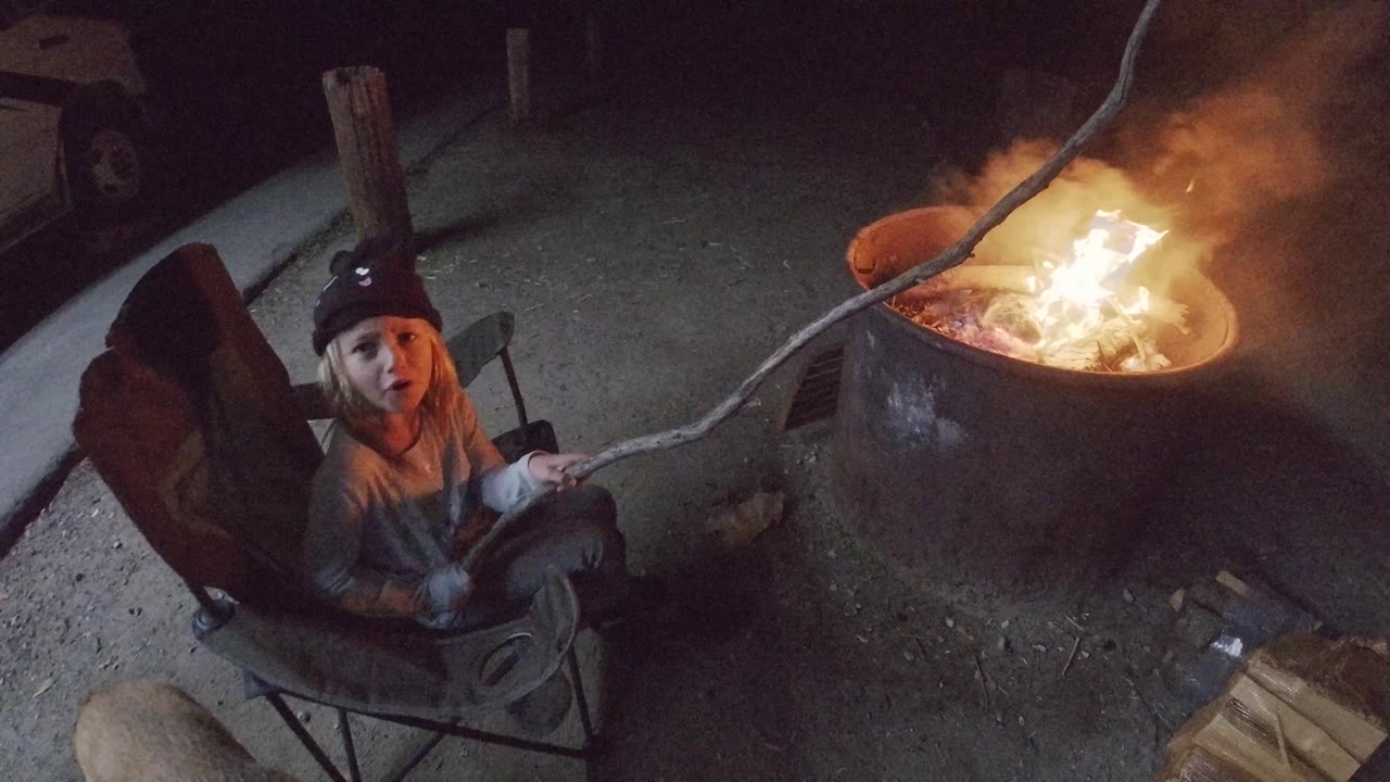 At 0:02 in San Clemente, the child remains seated on the left while smoke thickens above the round fire pit and the stick crosses over the flames.