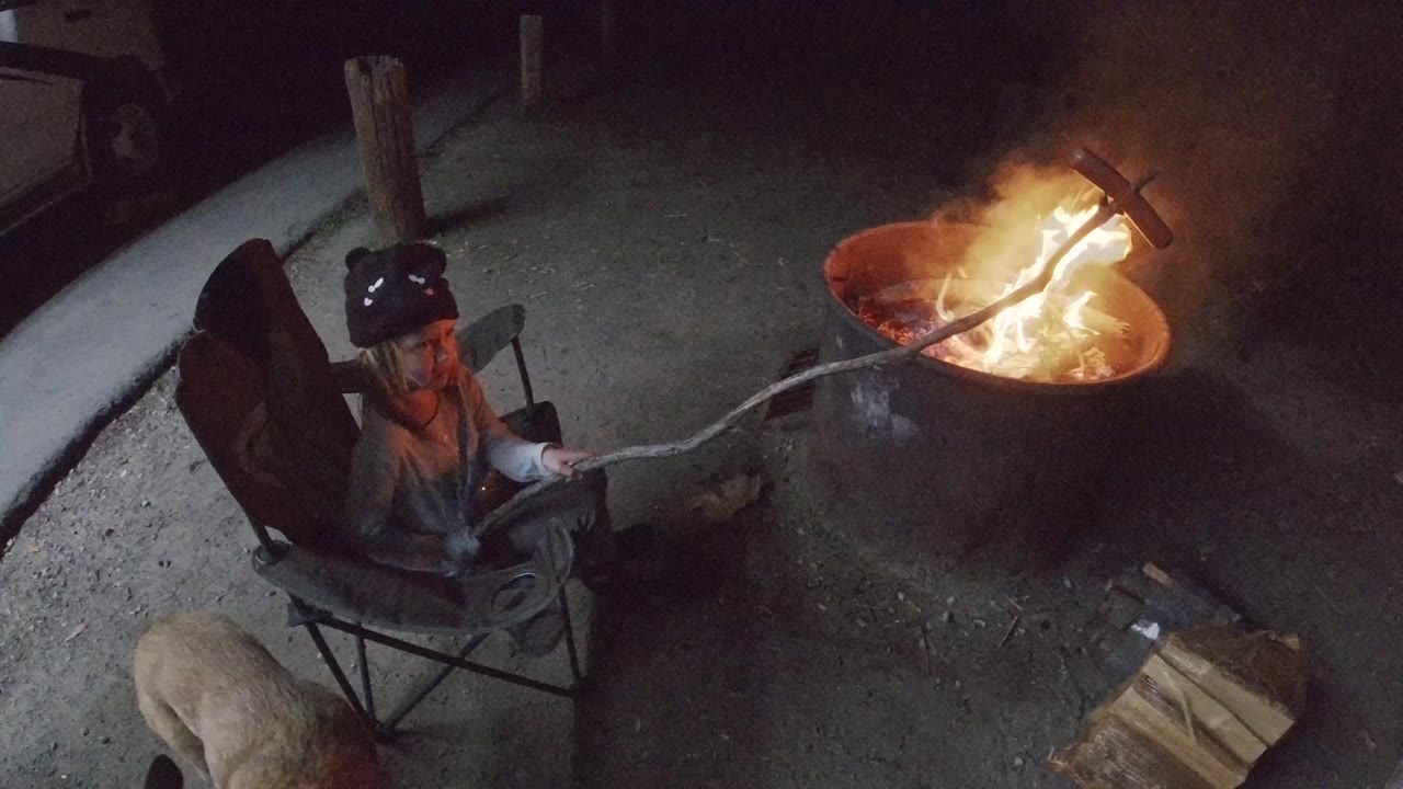 At 0:00 in San Clemente, California, a woman in a dark beanie sits in a folding chair holding a long stick toward a bright fire pit, with a light-colored dog partly visible below.