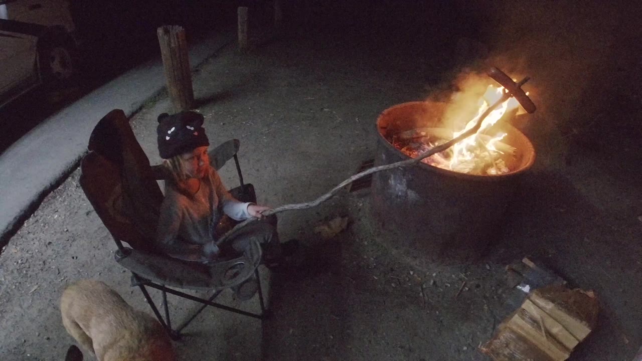 At 0:01, the woman remains seated in the chair as the long stick extends diagonally into the San Clemente fire pit, where flames brighten the upper right of the frame.