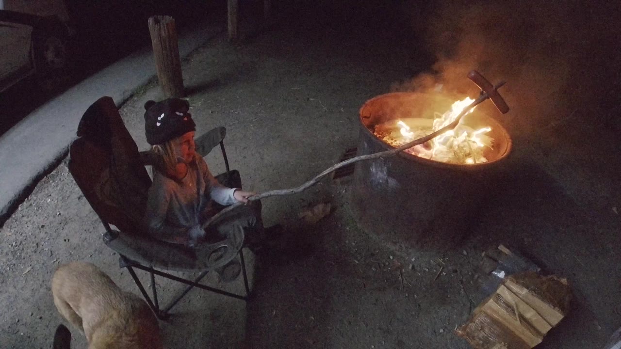At 0:02 in San Clemente, the fire flares higher and smoke gathers above the metal pit while the seated woman keeps the roasting stick extended from the chair.