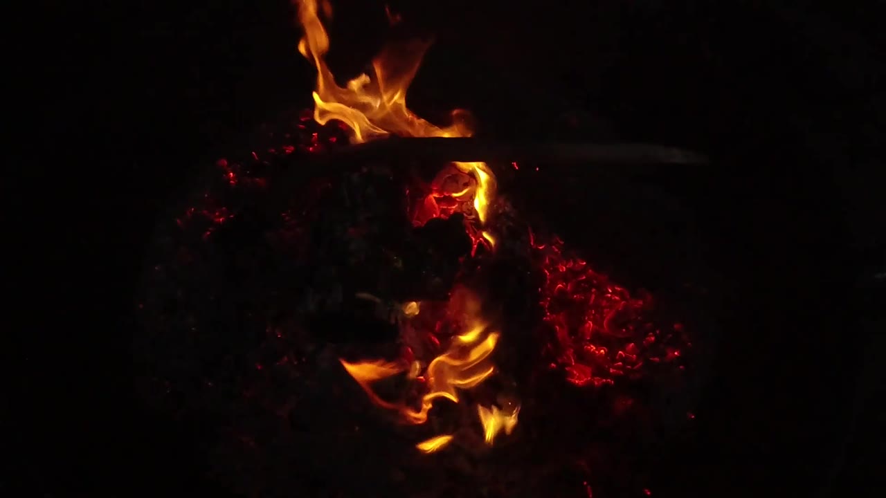Orange flames flare near the top and lower center of a dark campfire bed in San Clemente, California, with glowing red coals and no visible people.