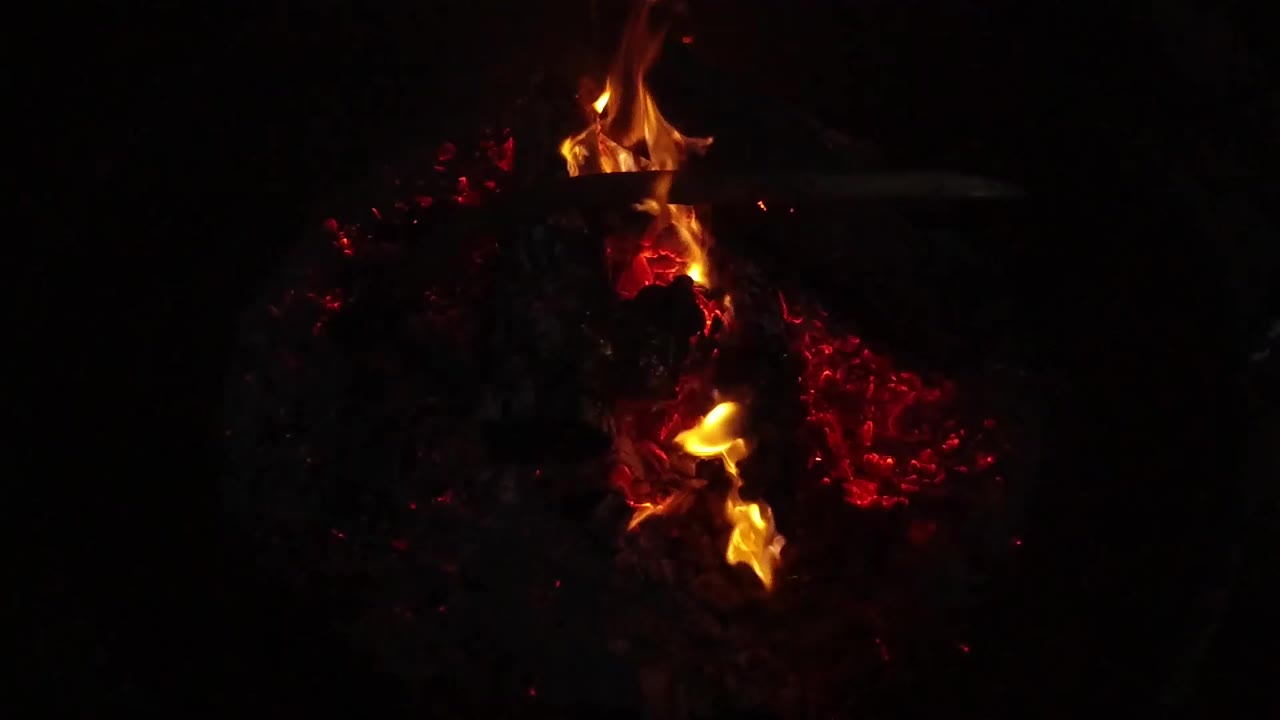 In San Clemente, California, small flames curl along charred logs while red embers glow on one side of the fire pit, with no visible people in this frame.