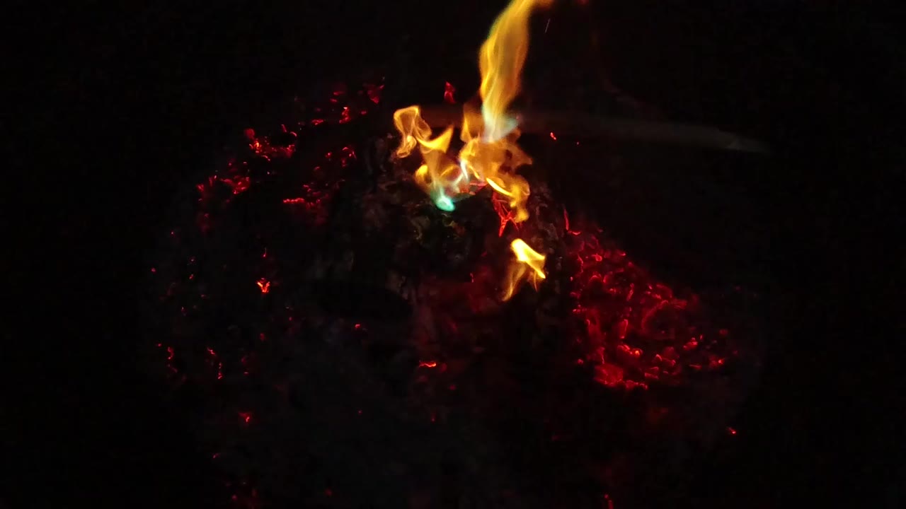A yellow flame with a small blue-green base burns over red embers in San Clemente, California, with the surrounding frame nearly black and no visible people.