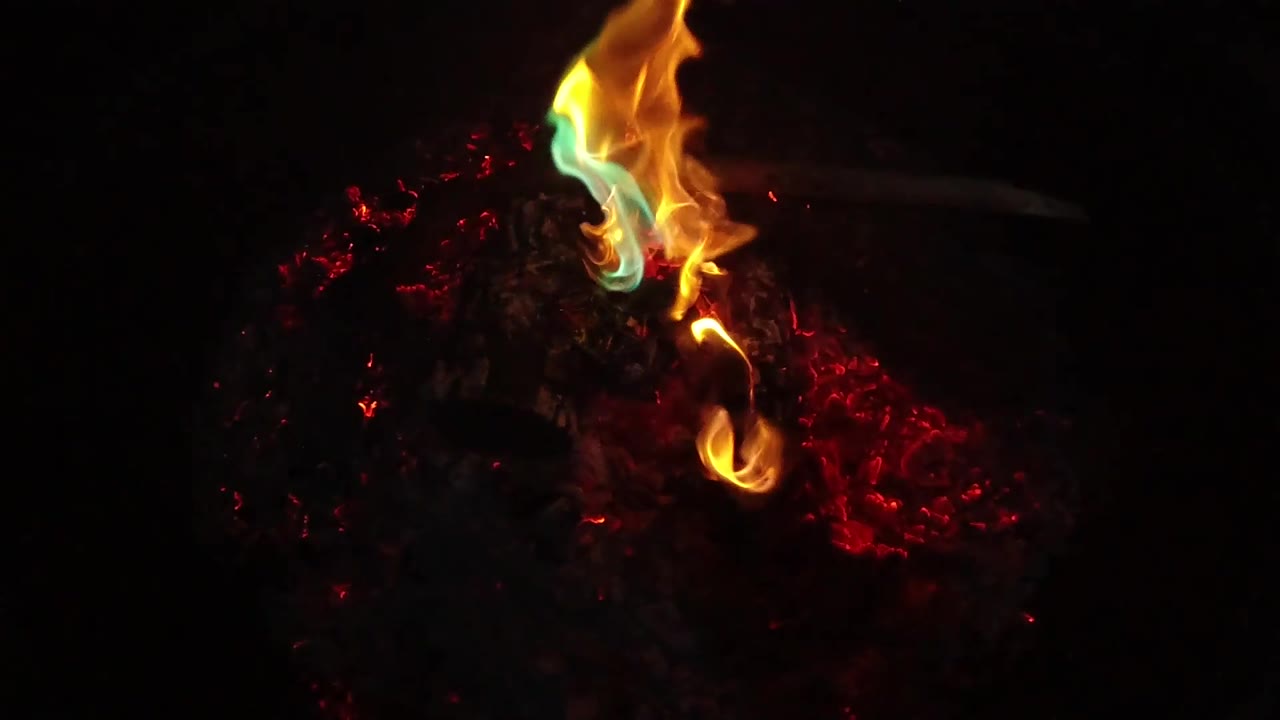 A taller yellow flame with a blue-green edge burns over the dark material in San Clemente, California, with red embers spread to the right and no visible people.