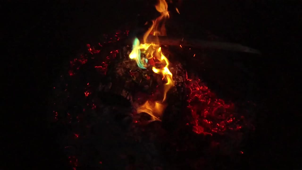 At the opening moment, orange flames and small green-blue tips burn over a charred log and red embers in San Clemente, California, with no visible people.