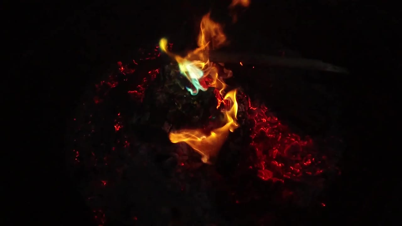 At two seconds, the orange flame bends around the front of the charred log with a green-blue flame patch near the top and red coals glowing at right, with no visible people.