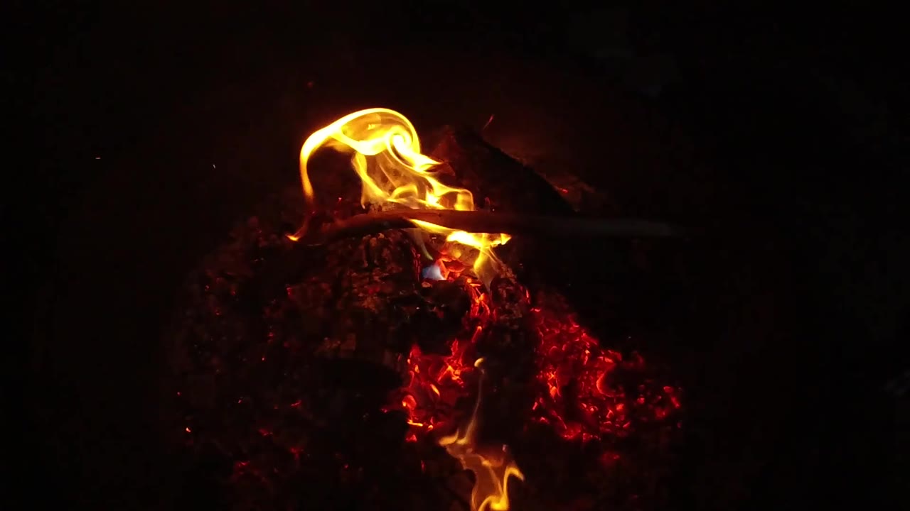 A loop of yellow-orange flame arcs across the top of the ember pile in San Clemente, California, while the surrounding fire pit stays very dark and no people are visible.