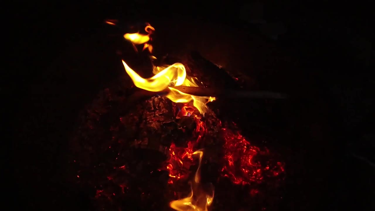 A broad orange flame spreads low across the left side of the red embers in the San Clemente fire pit, with no visible people in the dark frame.