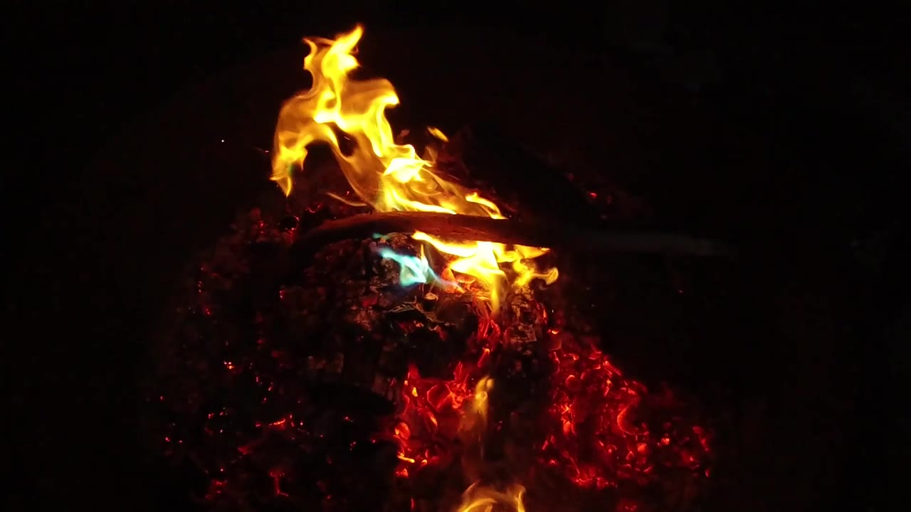 A taller yellow flame rises from the left side of the red ember bed in the San Clemente, California footage, with the background mostly black and no visible people.