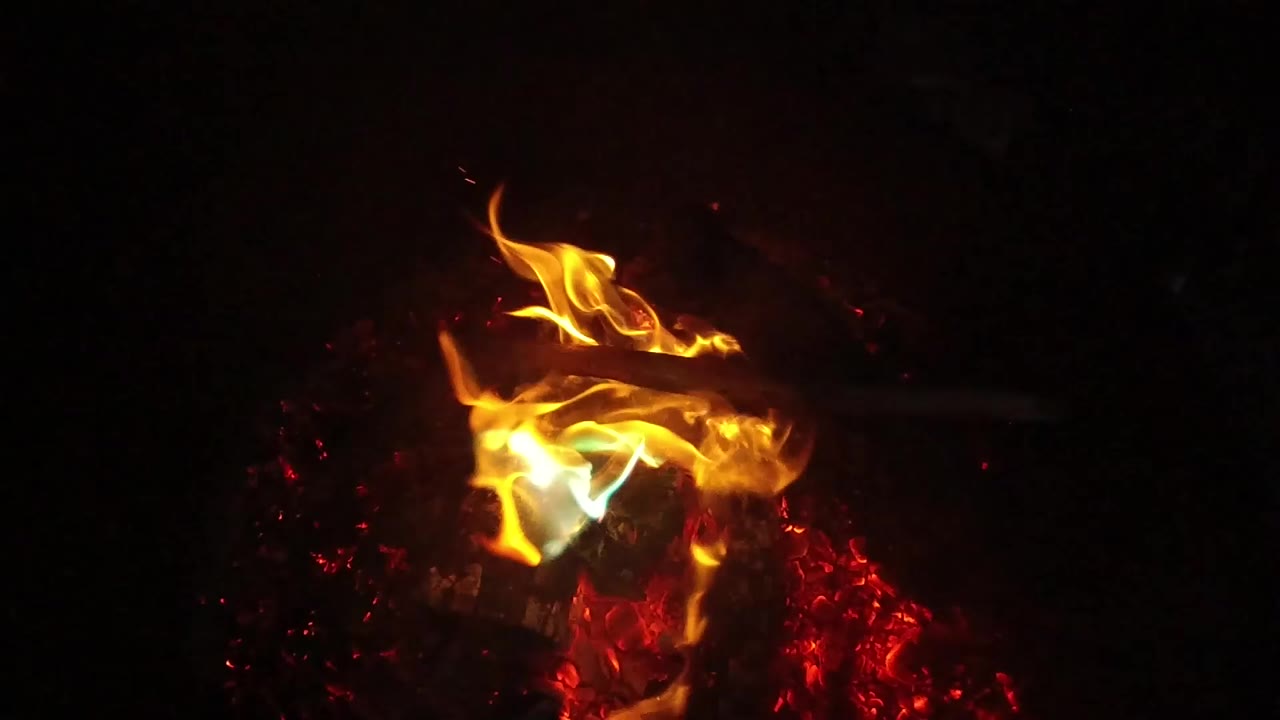 Close view of orange flames and a small blue-green flame burning over charred wood and red embers in San Clemente, California, with no visible people.