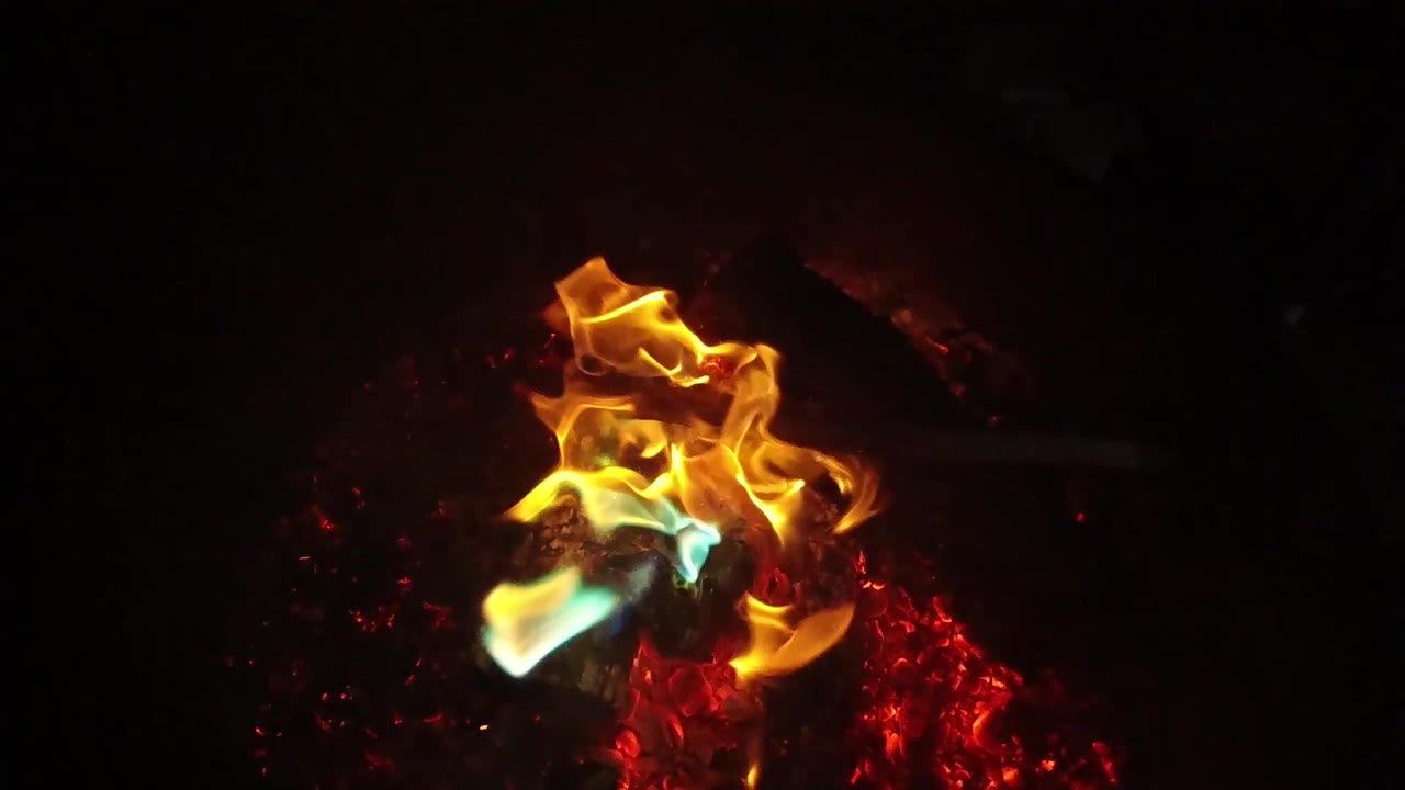 A taller orange flame lifts above a central burned log with blue-green color near the base and red embers to the side, with no visible people.