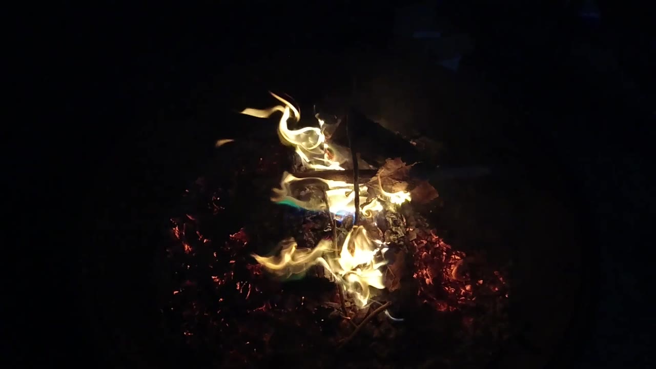 At 0:02, the flame shifts toward the center of the San Clemente fire pit, lighting red embers while most of the frame remains in deep shadow and no people are visible.