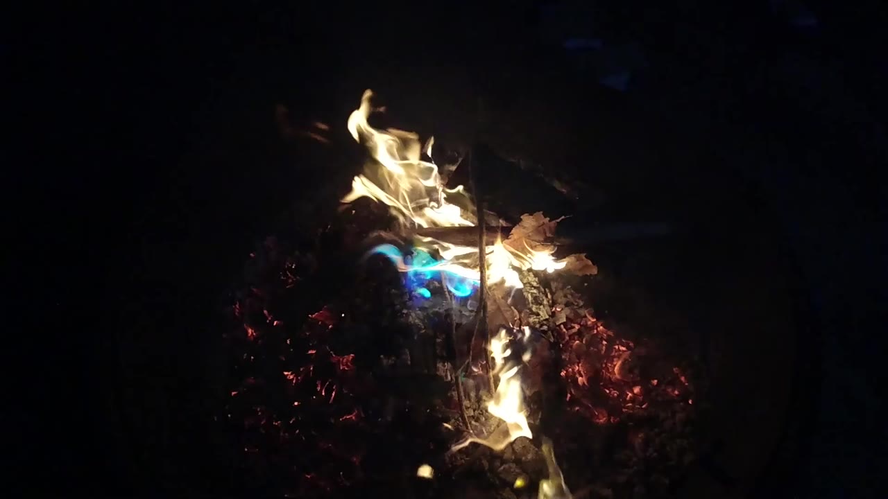 Tall yellow-white flames curl upward from charred logs, with a small blue flame visible near the center and no visible people.