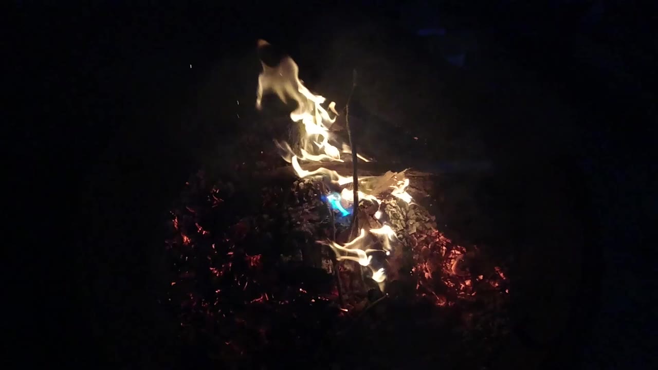 Bright yellow flames rise from red coals in a dark fire pit in San Clemente, California, with a small blue flame patch and no visible people.