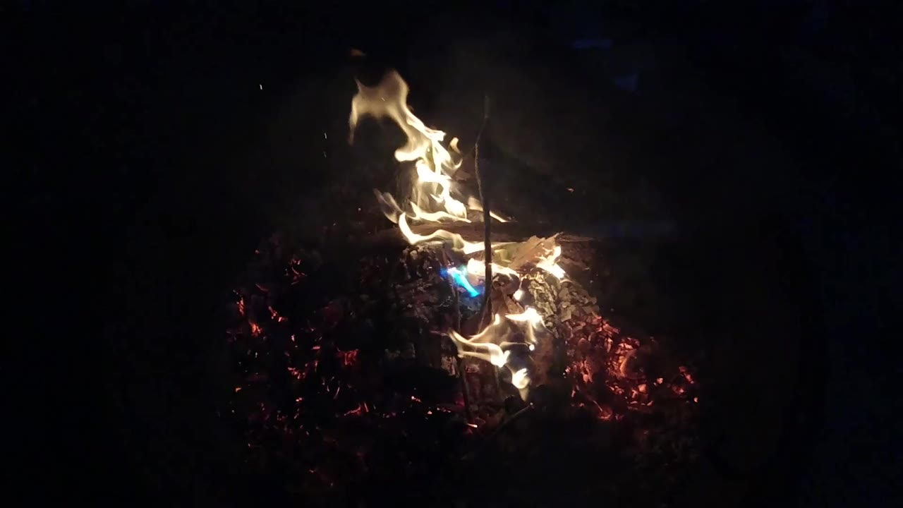A taller flame column curls above glowing red coals in the San Clemente fire pit, with the surrounding frame nearly black and no visible people.