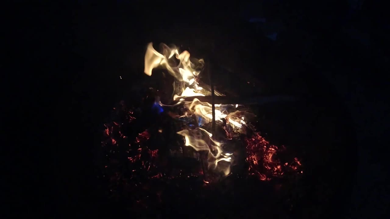 Curled yellow-white flames lift from the ember bed in the San Clemente fire pit, with red coals below and no visible people in the dark surroundings.