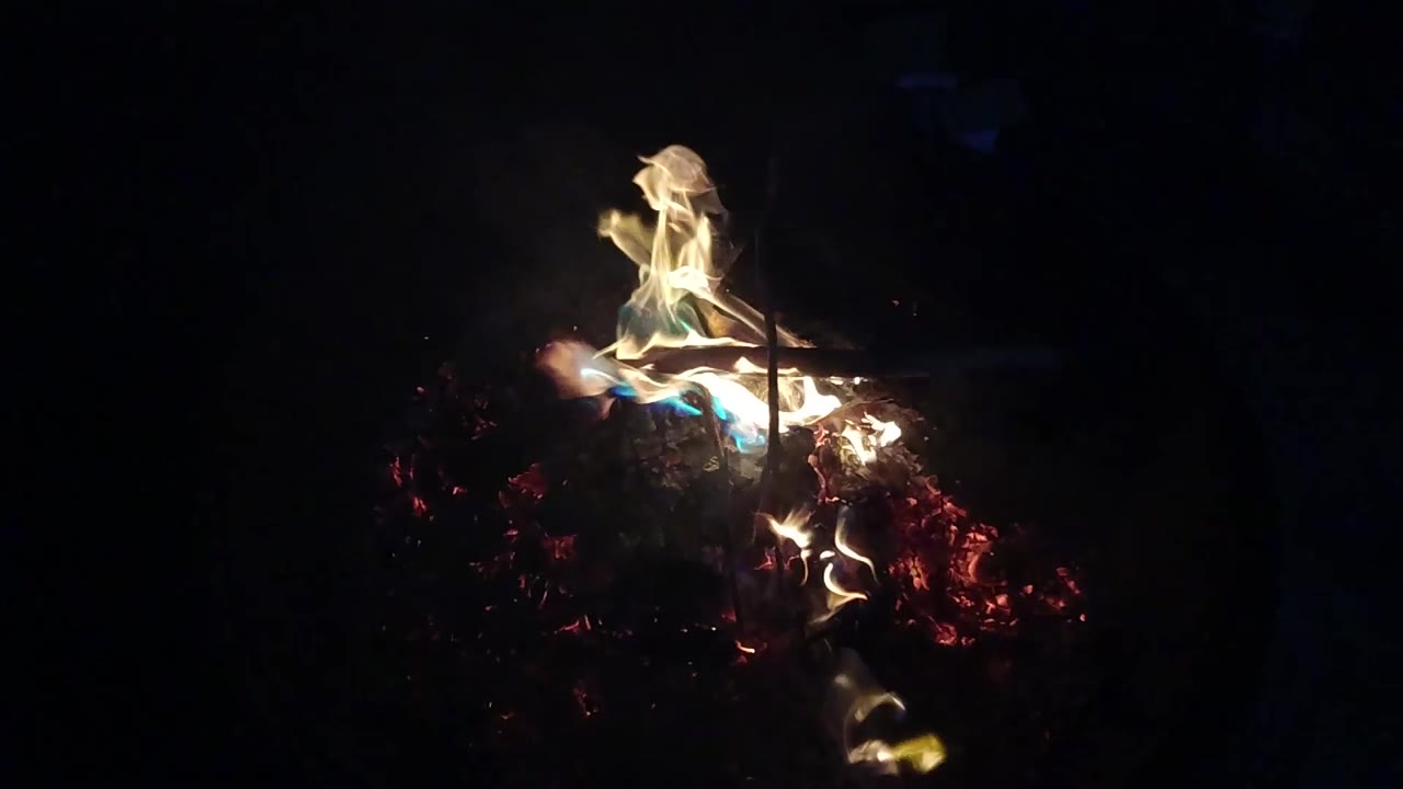 Flames rise from the upper edge of the wood pile in the San Clemente fire pit, with red embers glowing below and no visible people.