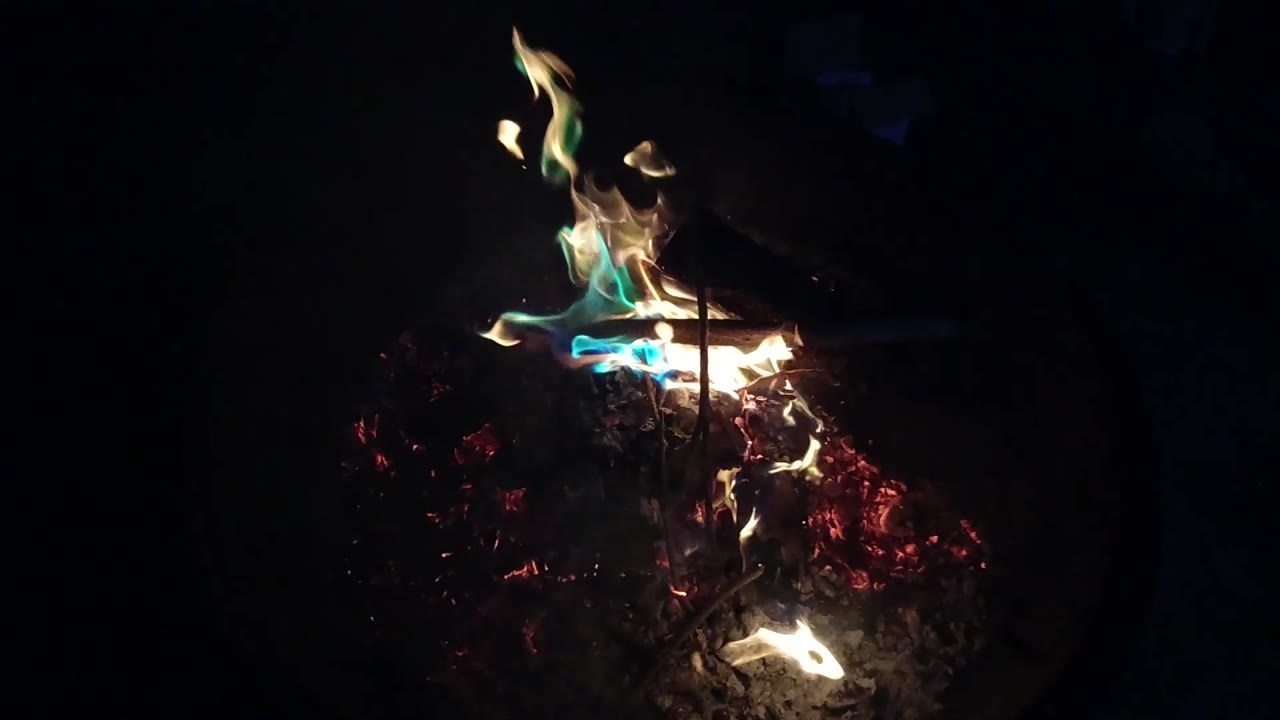 Blue-green and yellow flames burn across the left side of the wood pile in the San Clemente night fire pit, with no visible people.