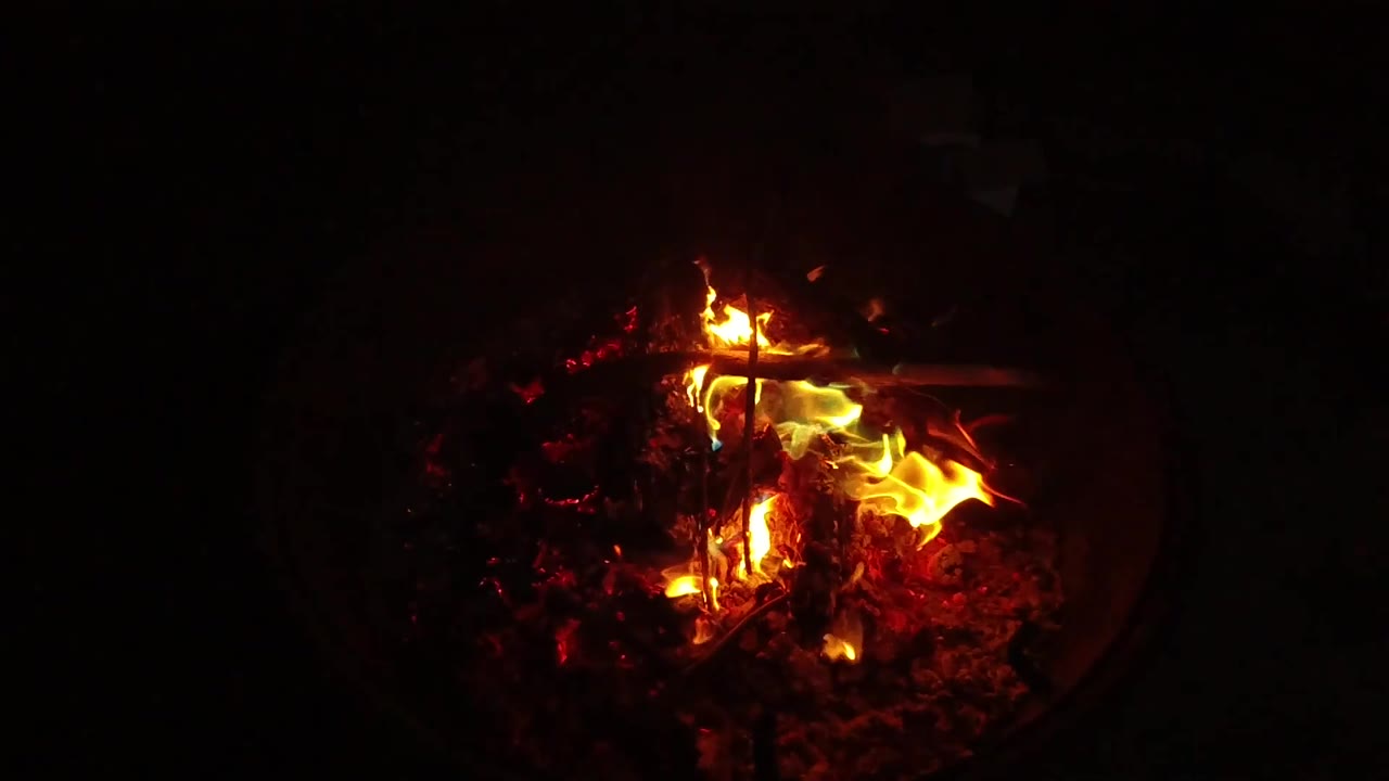At 0:02, yellow-orange flames lean right over a bed of red coals and blackened wood in the contained fire, with no visible people.