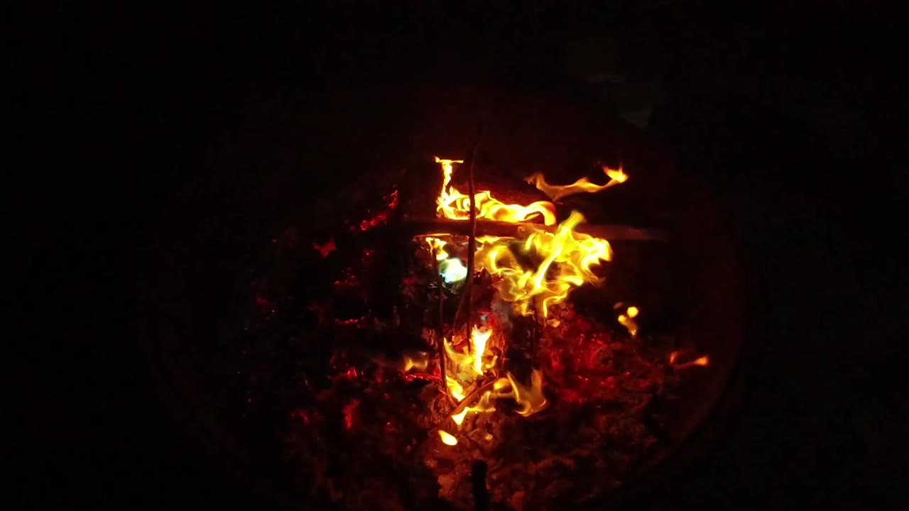 At 0:01, the flame front spreads wider across the embers in the dark fire pit, with a bright yellow tongue reaching to the right and no visible people.