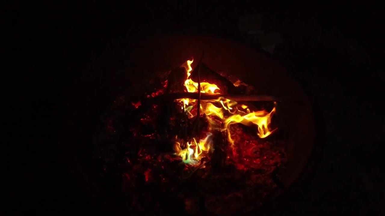 At 0:03, a taller yellow flame lifts from the left side of the ember bed inside the dark round fire pit, with no visible people in the surrounding black frame.