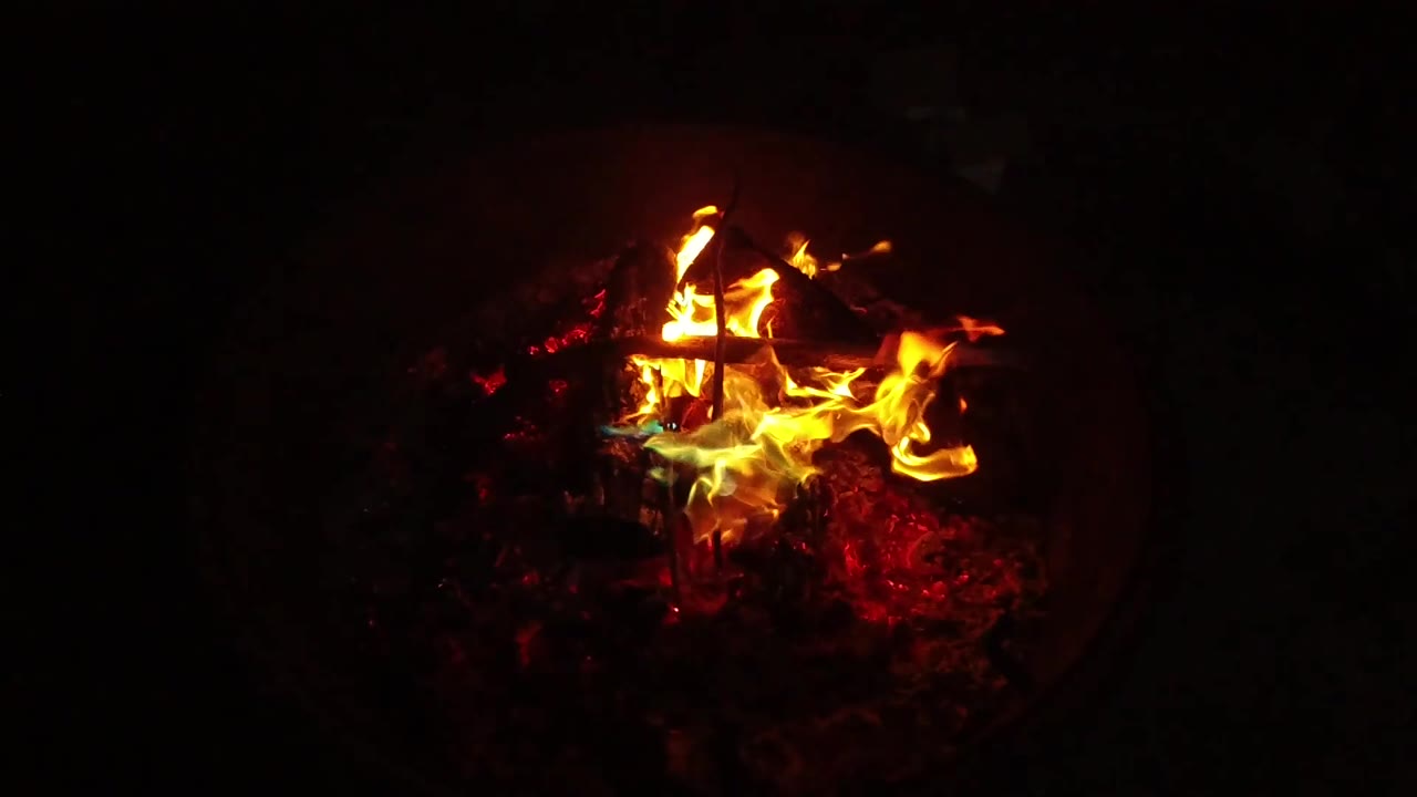 At 0:02, the fire flares wider to the right side of the crossed sticks, lighting red coals inside the dark container with no visible people.