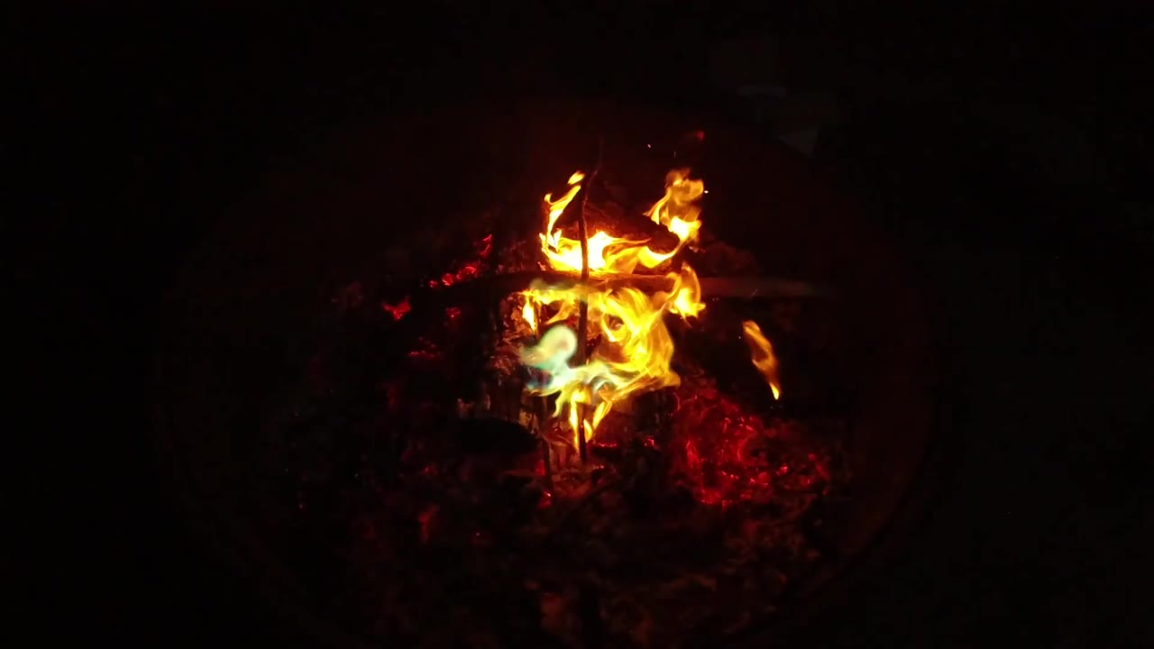 At 0:03, lower flames curl around the sticks while red embers fill the lower part of the container, and no people are visible in the dark frame.
