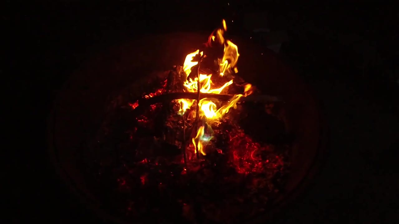 A bright flame burns inside a dark round fire container in San Clemente, California, with red coals underneath, a metal grate shape across the center, and no visible people.