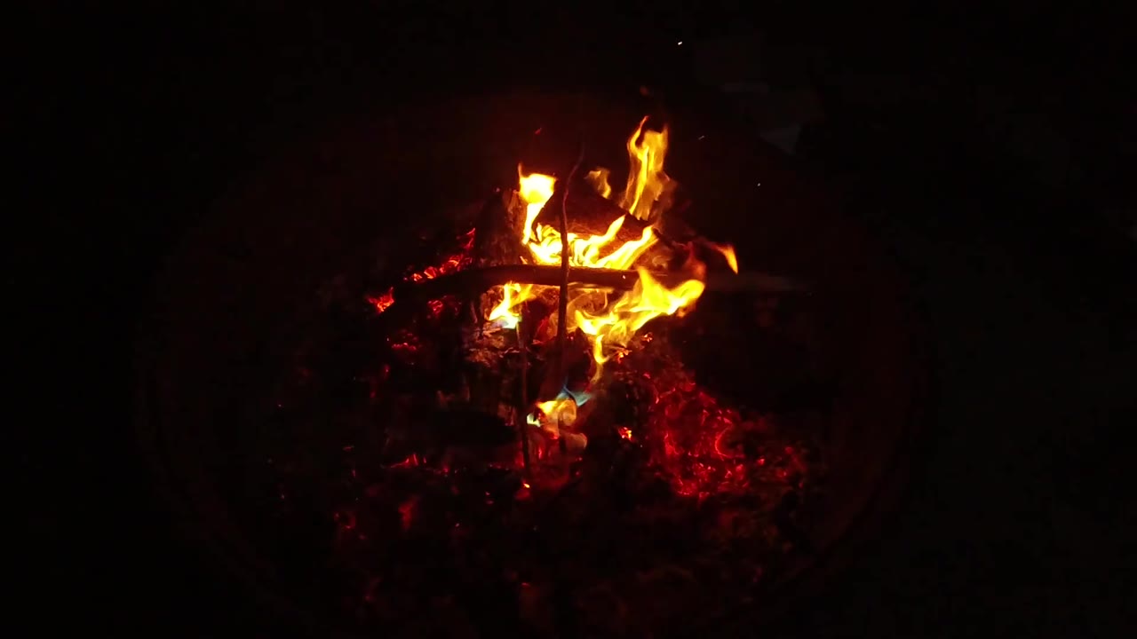 The flame spreads lower across the ember bed in San Clemente, California, lighting a dark metal crossbar while the rest of the frame stays black and no people are visible.