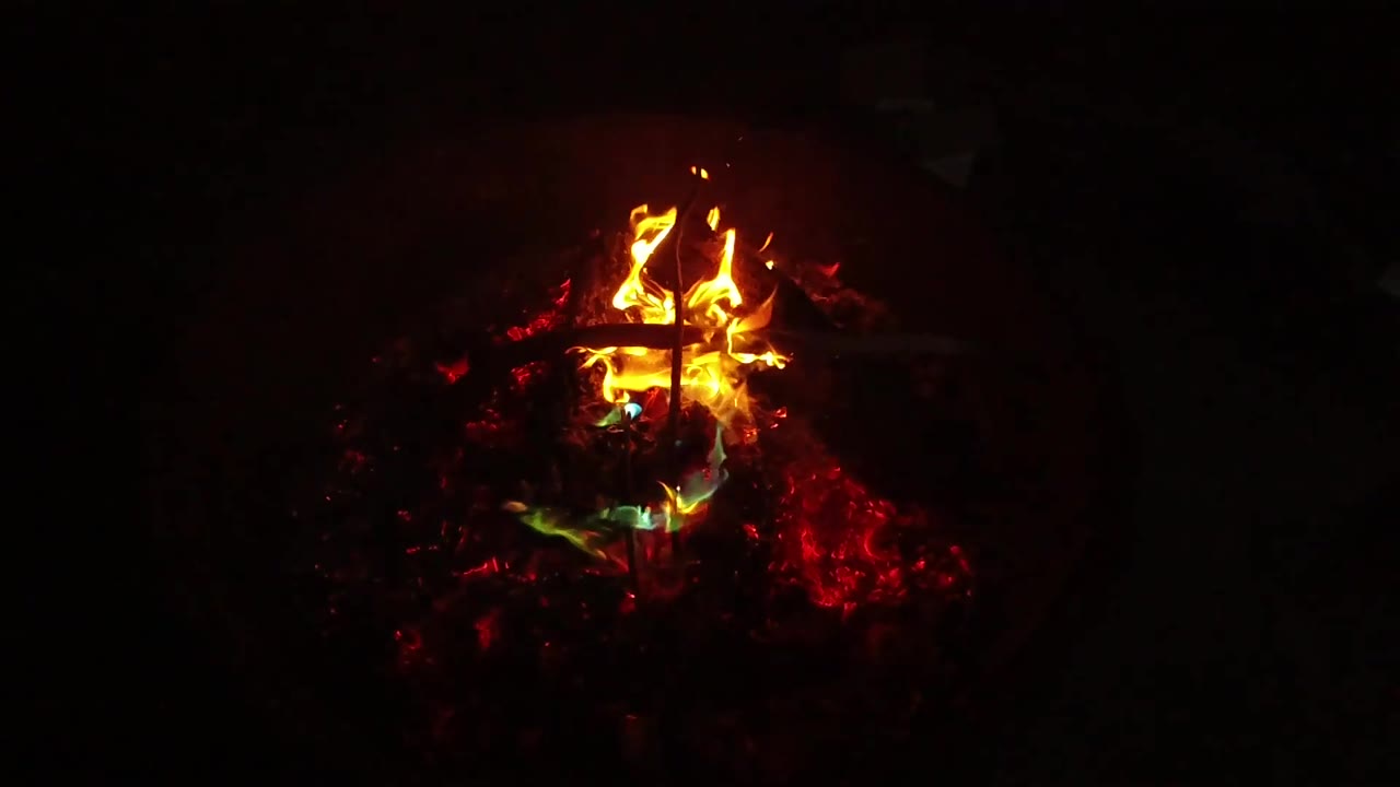 Small fire burns low over red coals in darkness.