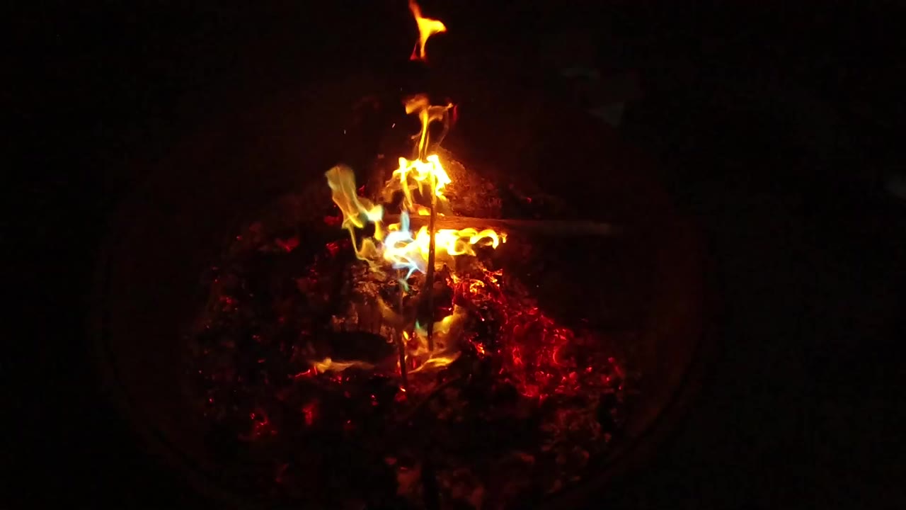 At 0:01, brighter flames curl upward from the ember bed, with the round fire pit still mostly surrounded by black shadow and no visible people.