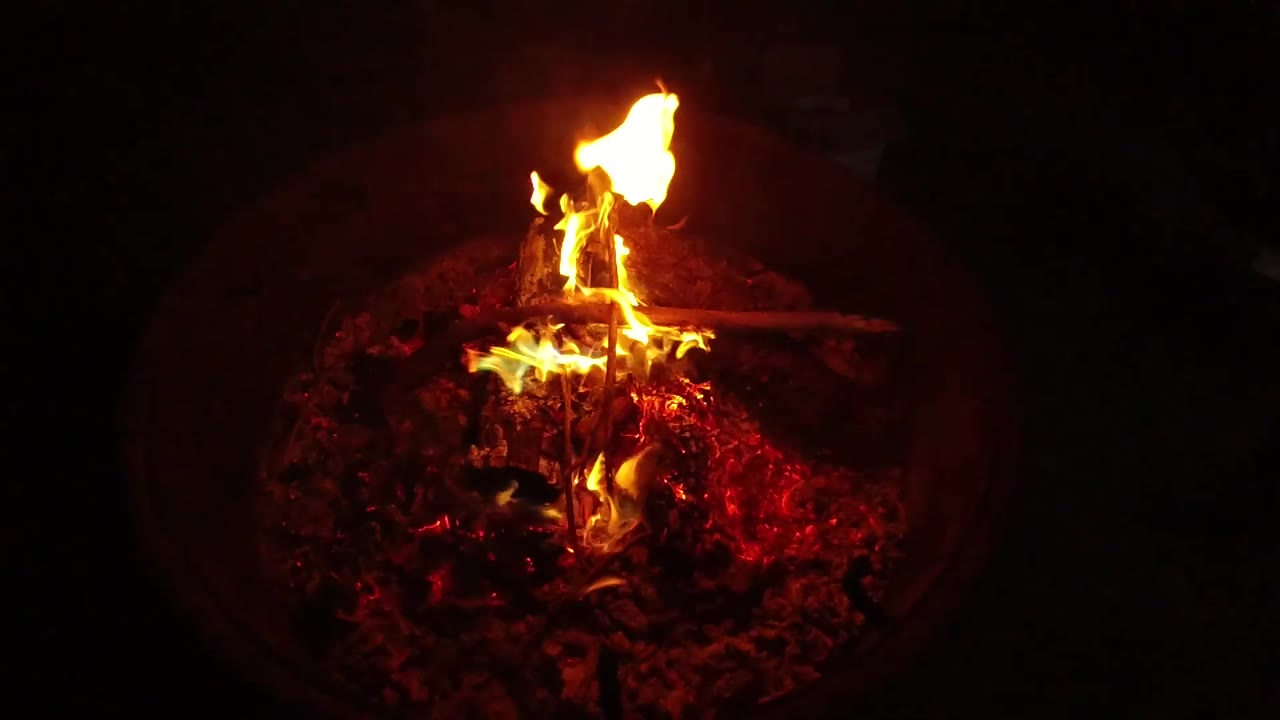 At 0:02, a taller flame lifts from the upper side of the wood while the ember bed glows red inside the circular fire pit, with no visible people.
