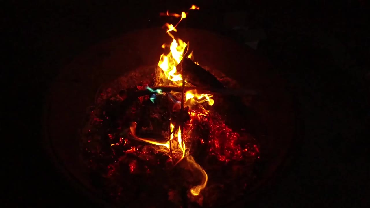A tight high-angle view of a small barbecue fire in San Clemente, California, with orange flames rising above red embers and no visible people.