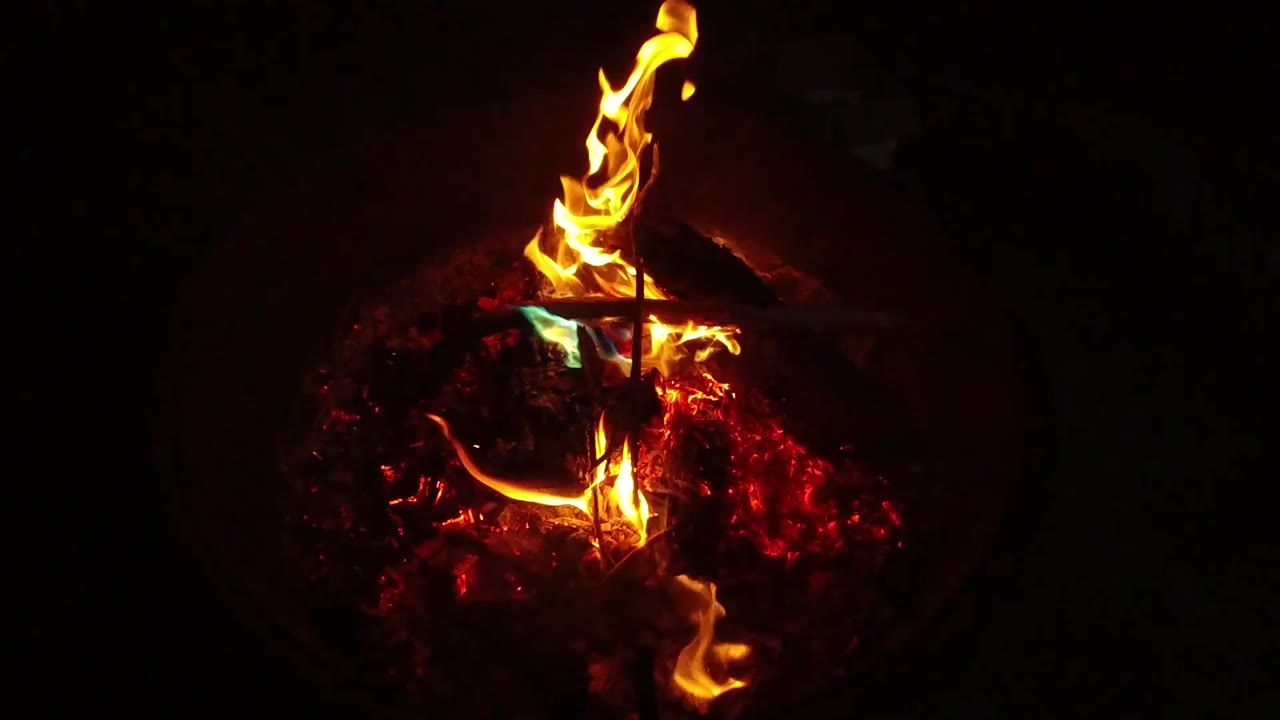 A curling flame reaches upward from the barbecue fire while red embers glow beneath it in the surrounding darkness, with no visible people.
