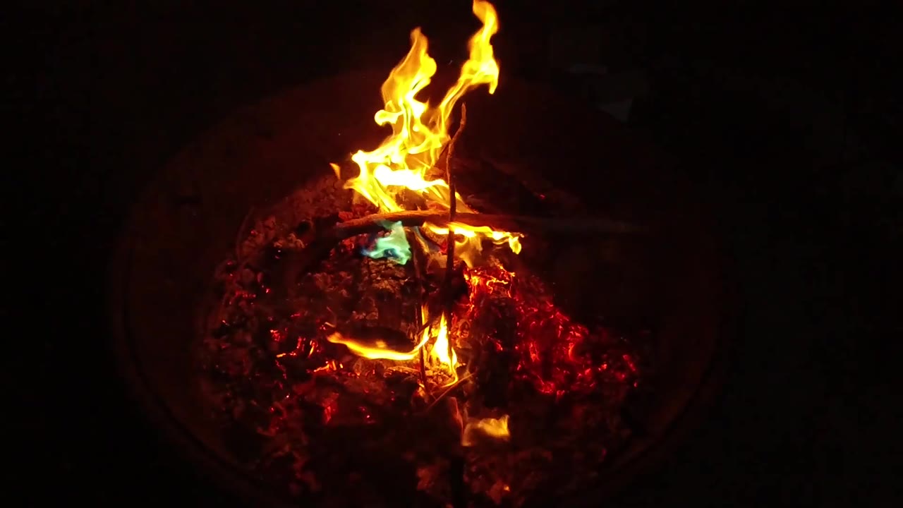 A bright orange flame flares above the ember pile, with the fire pit edge barely visible in the dark and no visible people.