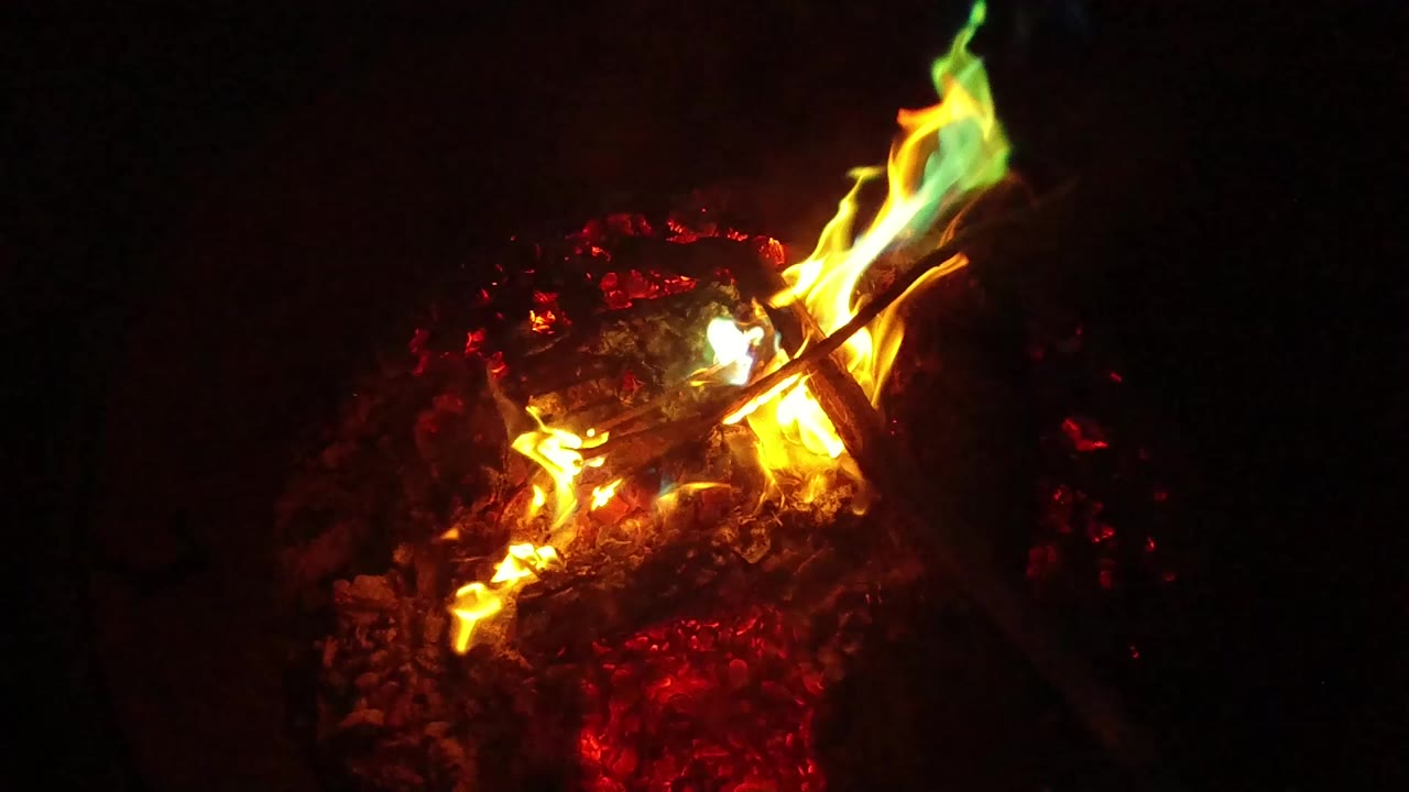 A controlled bonfire in San Clemente, California begins with orange flames rising from glowing red embers, a charred stick crossing the fire, and no visible people.