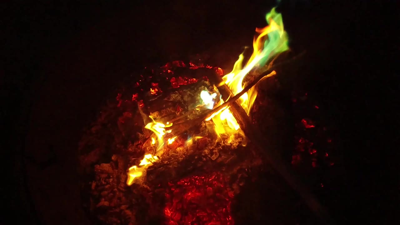 Orange and yellow flames stretch diagonally over the ember bed in San Clemente, California, with the stick still crossing the frame and no visible people.