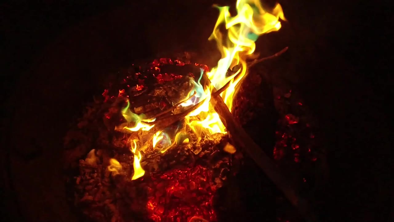 At 0:01, a taller flame leans upward over glowing red coals and crossed twigs in the San Clemente brazier, with no visible people.