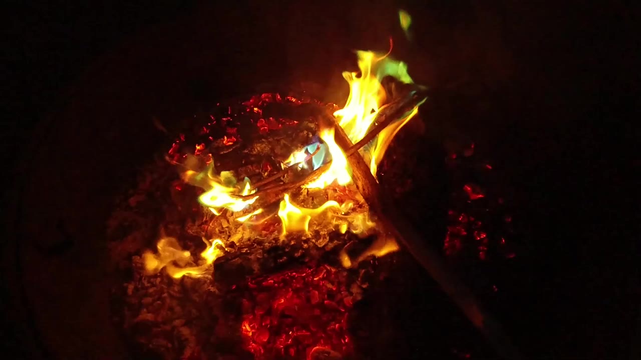 At 0:00, yellow flames burn across charred wood and red embers in San Clemente, California, with a dark stick angled through the frame and no visible people.