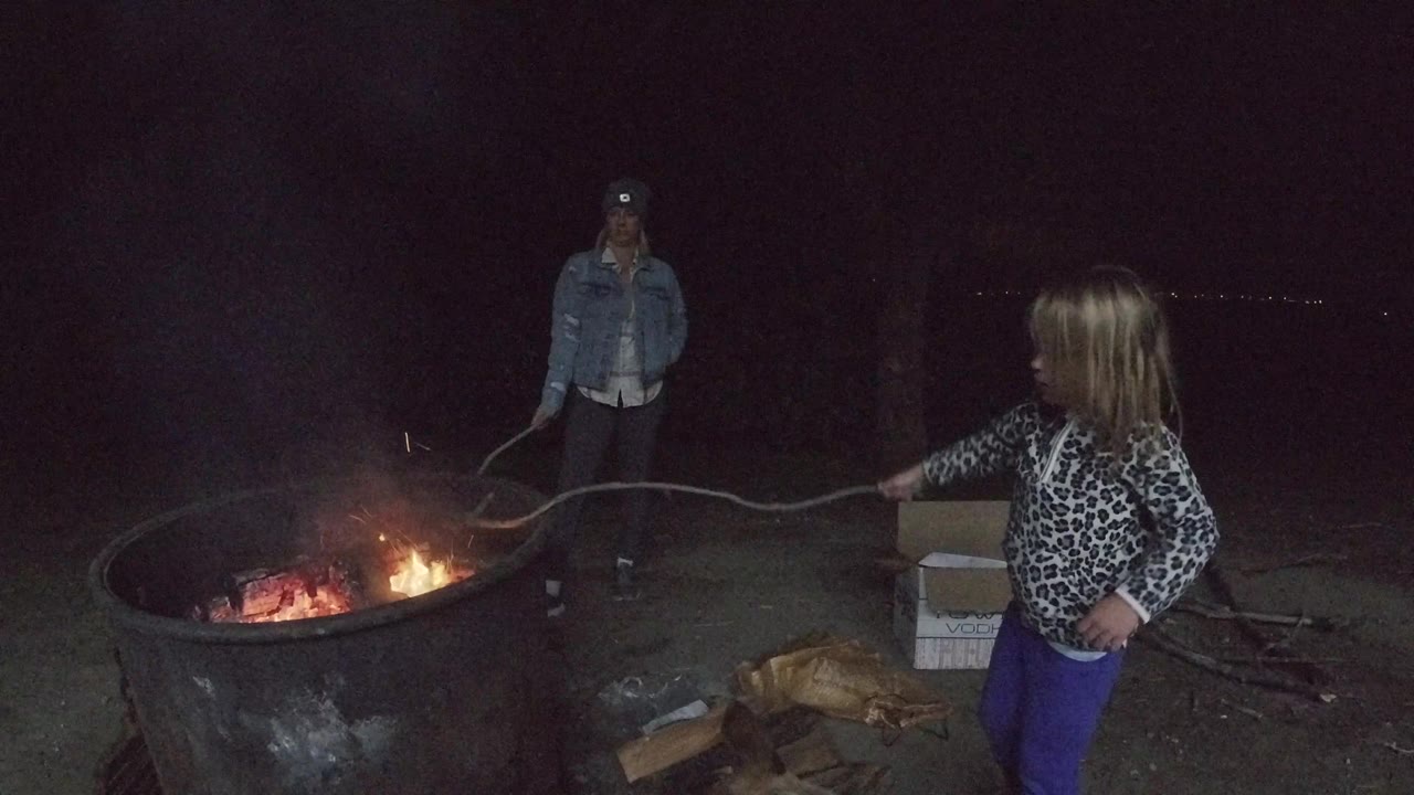 In San Clemente, California, the girl stands to the right of the metal fire pit with the stick stretched across the frame and lower flames glowing inside the bowl.