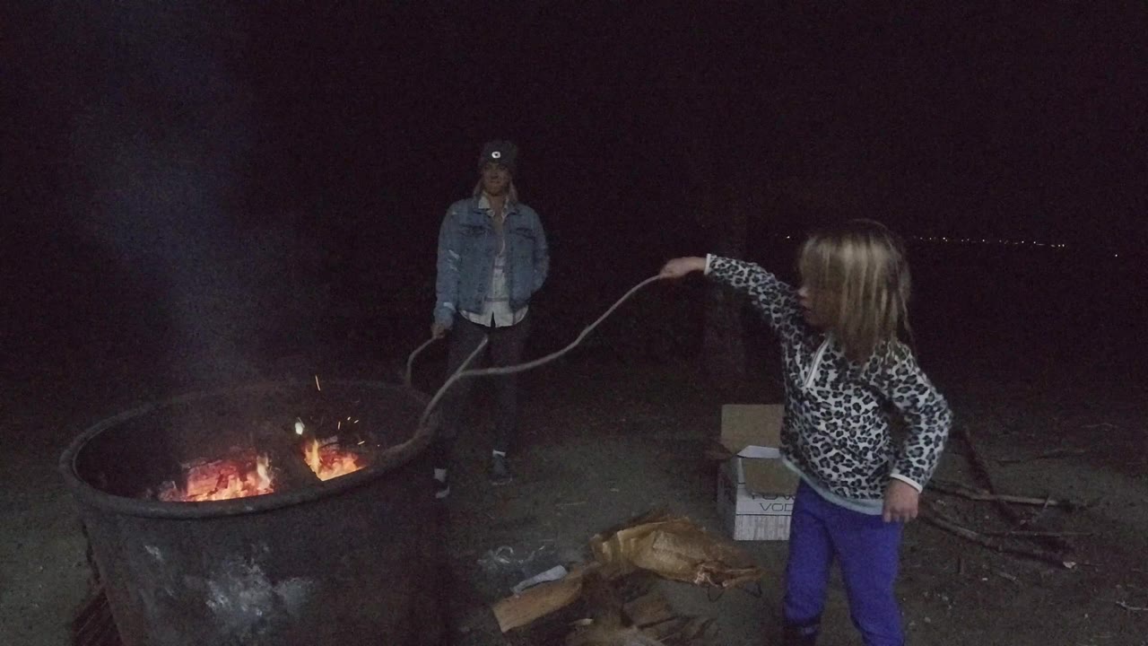 Girl leans toward a campfire with a stick while smoke rises in the dark.