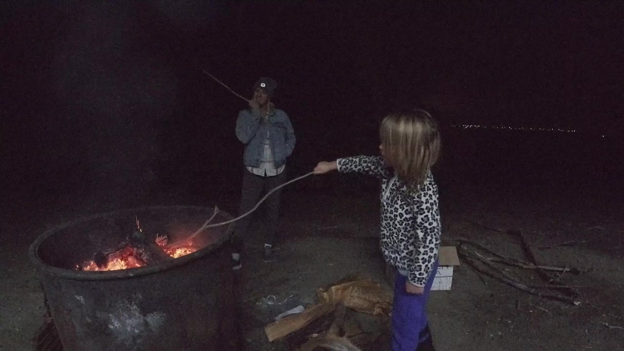 In San Clemente, California, a child in purple pants extends a long stick toward a glowing metal fire ring while an adult man stands in the dark behind the flames.