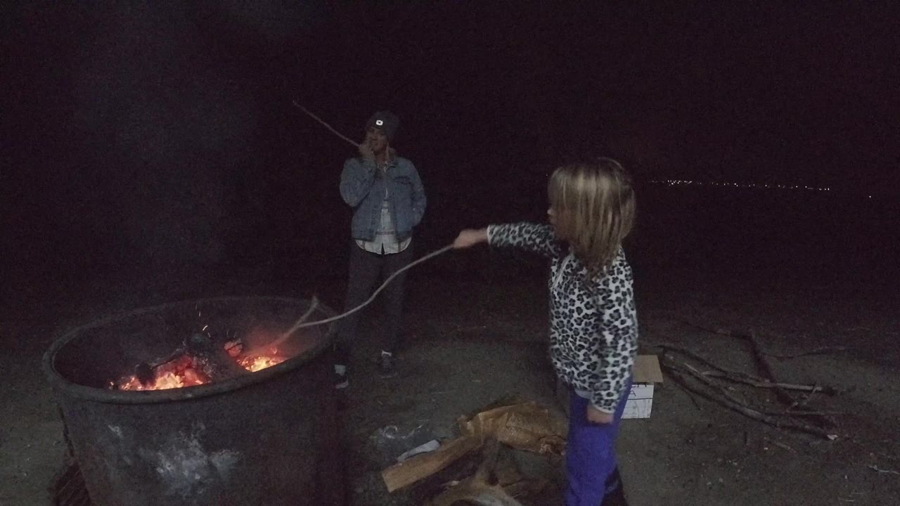 Child keeps a stick over the fire ring as an adult watches from behind the flames.