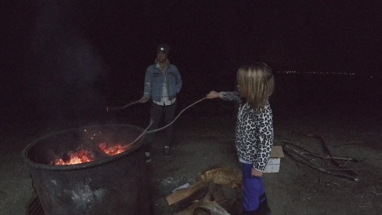 A child at the right edge and an adult man behind the fire ring face each other in San Clemente, California, as smoke rises from the glowing coals.