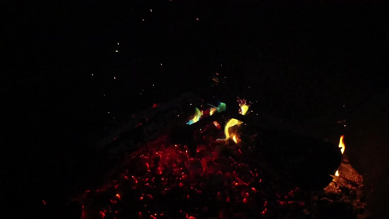 Burning logs sit over red coals in a dark San Clemente fire pit scene, with a small flame at right and no visible people.