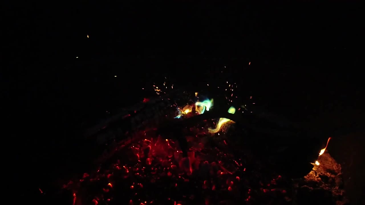 A low fire pit in San Clemente shows red coals under dark logs, with a bright flame licking up on the right and no visible people.