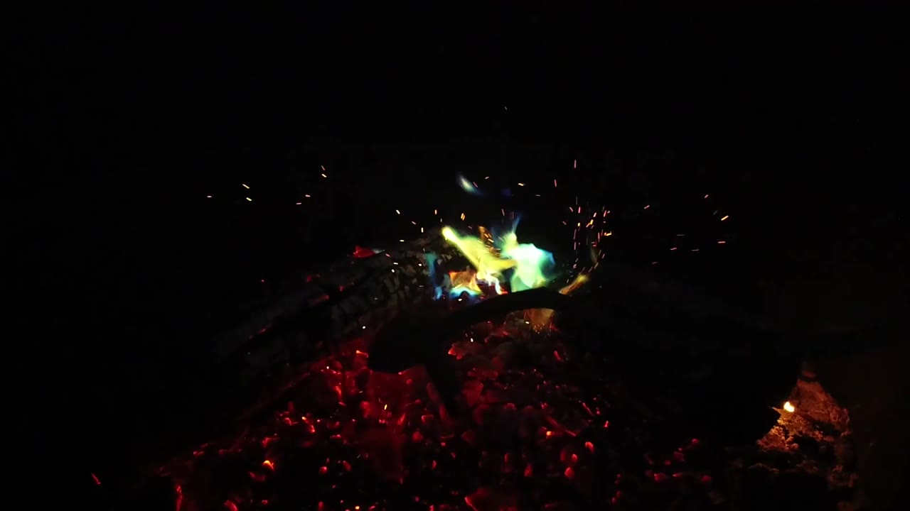 A green-blue flame flares over the left side of the burning logs in the San Clemente fire pit, with red coals below and no visible people.