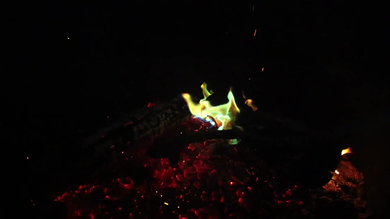 At 0:02, the green-yellow flame leans across the top of the ember pile with scattered sparks above the dark fireplace area, with no visible people.